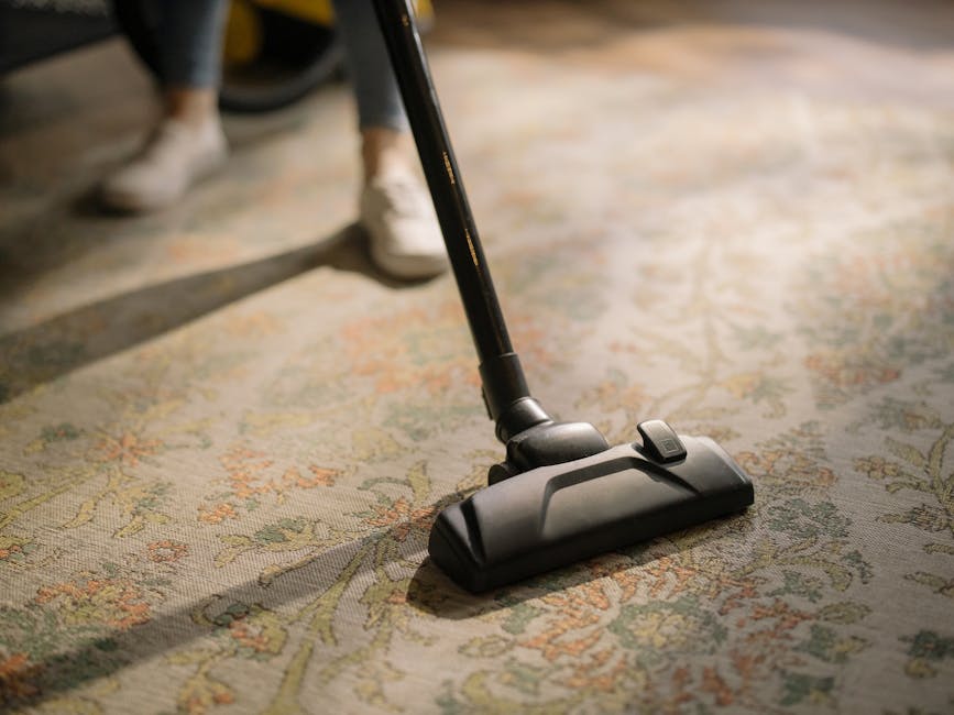 A person operating a yellow and black carpet cleaning vacuum cleaner on a patterned beige and green area rug in a residential living room. The individual is wearing light-colored shoes and jeans, with